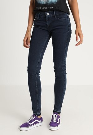 Jeans ajustados de mezclilla color azul oscuro, con una cintura de altura media, cinco bolsillos y un diseño limpio. Llevados con zapatillas moradas.