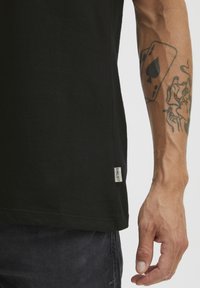 Fekete pamut póló rövid ujjakkal, enyhén karcsúsított szabással és egy kis logófával az alján. Látható tetoválás a karon.