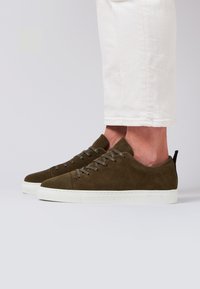 Zapatillas de ante verde con diseño de cordones, suela de goma blanca y tira en el talón negra. Detalle de costura visible a lo largo del borde de la suela.
