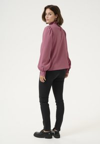 Blouse bordeaux avec des manches bouffantes et un col haut à volants, associée à un jean skinny noir et des chaussures à plate-forme noires, vue de dos.