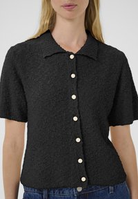 Femme portant un haut noir à manches courtes texturé avec des boutons en perles et un jean en denim bleu sur fond uni.