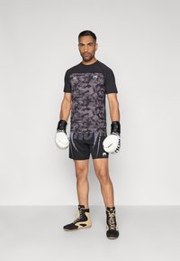 Mustas ja hallis kamuflaaživärvides treeningpluus, mustad lühikesed püksid aktsentidega, poksikindad ning mustad ja beežid kõrged tossud, seisab ühevärvilise tausta ees.