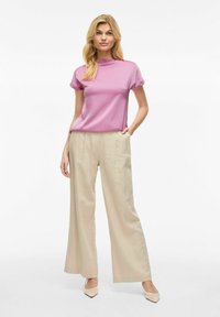 Top corto rosa chiaro a maniche corte abbinato a pantaloni beige a gamba larga e tacchi chiari, con una texture liscia e un fit moderno.