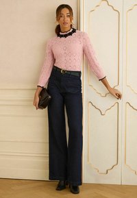 Pulover roz deschis tricotat cu modele în formă de diamant și dantelă neagră la margine, asortat cu blugi evazați de culoare albastru închis și cizme negre cu toc. Geantă maro.