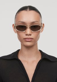 Ochelari de soare cu o formă rectangulară, rame transparente bej și lentile întunecate, având un design elegant cu accente minime.