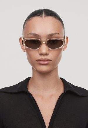 Lunettes de soleil de forme rectangulaire, avec des montures transparentes beige et des verres teints foncés, présentant un design épuré avec peu d'accents.