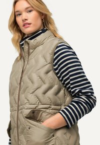 Beżowa pikowana kamizelka z wysokim kołnierzem i brązowym zamkiem, noszona na granatowej i białej w paski koszuli z długim rękawem. Posiada boczne kieszenie.