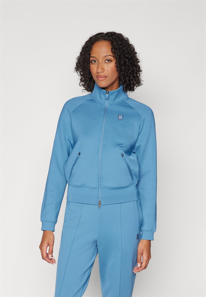 Blaues Zip-Jacke mit hohem Kragen, ausgestattet mit zwei vorderen Reißverschlusstaschen und gerippten Bündchen. Weiches Material mit glatter Textur.