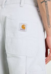 Witte katoenen cargobroek met een grote zijzak en een gestikte Carhartt-logo patch in oranje en bruin. Versterkte stiksels zichtbaar.