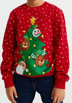 Barn iført rød julesweater med dekoreret grøn træ med ornamenter formet som snemænd, rensdyr, honningkage og julemandsansigter.