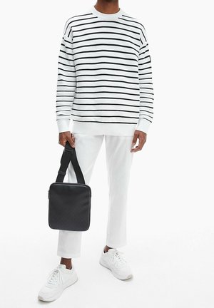 Personne portant un pantalon blanc, des baskets blanches et une chemise blanche à manches longues avec des rayures horizontales noires, tenant un sac à bandoulière carré noir.
