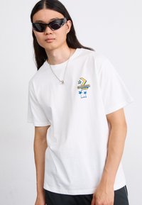 Hvid bomulds T-shirt med en lille flerfarvet grafik med stjerner og palmer. Modellen bærer sorte solbriller og en sølv halskæde.
