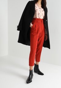 Cappotto nero, blouse con motivo floreale, pantaloni rossi a vita alta con cintura, stivali neri alla caviglia. Tessuti morbidi, vestibilità su misura, design moderno.
