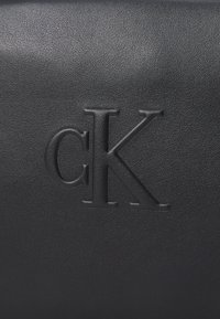 Svart texturerad läderväska med ett präglat "CK"-logotyp i en elegant design. Ytan är slät med en subtil kornfinish.