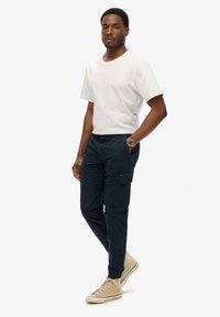 T-shirt blanc à manches courtes, pantalon cargo bleu marine avec poches latérales, et baskets beiges montantes. Design simple avec une coupe décontractée.