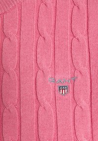 Maglione rosa lavorato a maglia con motivi a treccia, dotato di un logo GANT grigio e un emblema a scudo con strisce rosse e bianche.