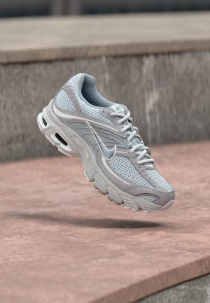 Gaiši pelēka un balta Nike skriešanas kurpe ar sieta augšdaļu un mīkstinātu zolīti, attēlota gaisā uz betona pakāpieniem fona.