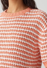 Acercamiento de una persona que lleva un suéter de punto con textura, a rayas naranjas y blancas, con cuello redondo y mangas cortas.