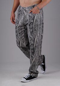 Man wearing black and white patterned pants and black sneakers with white laces, standing with one hand in pocket and one foot slightly raised.