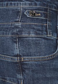 Detalle de tela de mezclilla azul oscuro con costuras visibles, un botón de metal y una pequeña tapa de bolsillo trasero en un par de jeans.