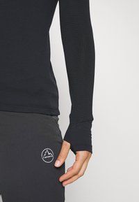 Svart långärmad termoshirt i texturerad väv, kroppsnära design och tumhål. Ett par mörka byxor med en liten vit logotyp på låret.