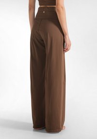 Donna che indossa pantaloni marroni a vita alta e gamba larga, in piedi a piedi nudi contro uno sfondo bianco semplice.