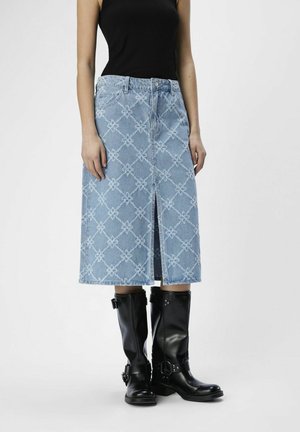 Femme portant une jupe en jean bleue à motifs mi-longue avec une fente à l'avant, un top noir sans manches et des bottes noires montantes avec boucle.