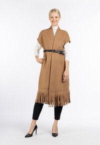 Brun frynset vest med bælte, lavet af et struktureret stof. Har et åben frontdesign og er stylet over en hvid langærmet top.