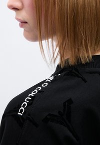 Μαύρο φούτερ με βελούδινη υφή, με κεντημένο λευκό όνομα εμπορικής επωνυμίας και περίπλοκα σχέδια κατά μήκος της ραφής στον ώμο.