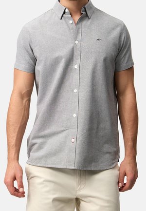 Mężczyzna w krótkim, jasnoszarym koszulce na guziki z małym czarnym logo na piersi, w jasnobeżowych spodniach.