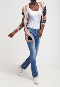 Lång, öppenstickad kofta med geometriska mönster i persika, marinblå och krämfärgad. Vit linne, blå jeans och vita sneakers fulländar outfiten.