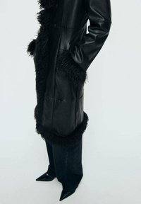 Palton din piele neagră cu detalii din blană sintetică, având două buzunare frontale, o siluetă lungă și margini rotunjite. Asortat cu blugi închis la culoare și pantofi cu vârful ascuțit.