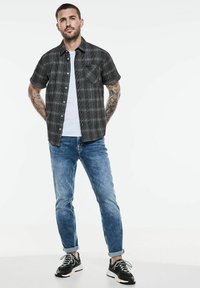 Krátký rukáv černé a šedé kostkované košile, bílý tričko pod tím, modré slim-fit džíny a černé tenisky s bílými akcenty.