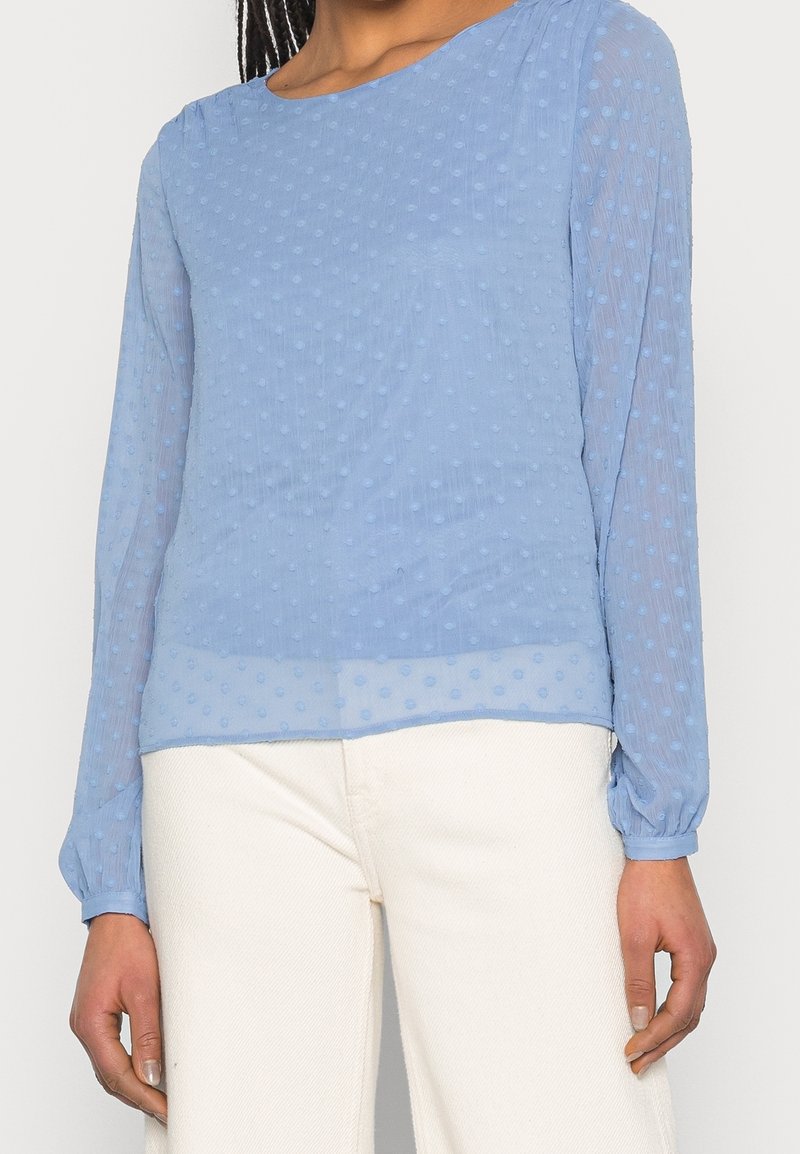 Femme portant un chemisier bleu clair, à manches longues, transparent texturé avec des points en relief, associé à un pantalon taille haute ivoire.