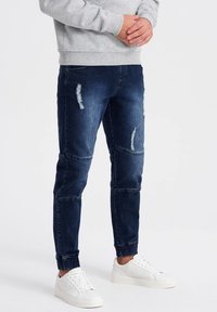 Jeans en denim bleu foncé avec des patches distendus et des jambes fuselées, portés avec des baskets blanches et un sweat-shirt gris.