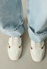 White sneakers with animal print accents, featuring a textured leather upper, round toe, and beige laces. Rolled-up light blue denim jeans.
