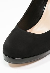 Černé nubukové lodičky na vysokém podpatku s kulatou špičkou a lesklým akcentem z lakované kůže podél podešve. Hladká textura a zúžený design.