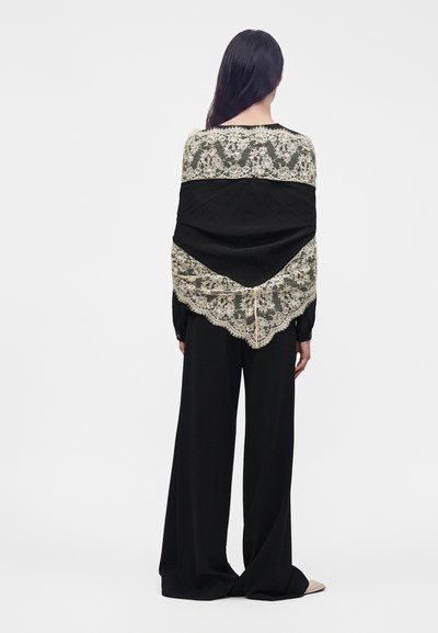 Personne aux longs cheveux foncés, de dos, portant un pantalon noir à jambes larges et un châle noir avec bordure en dentelle beige drapé sur les épaules.
