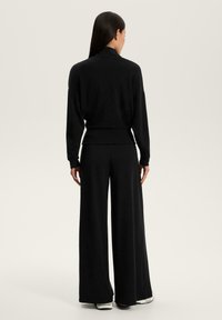 Femeie cu păr lung și închis la culoare, îmbrăcată cu un top negru cu mâneci lungi și pantaloni negri largi, stând cu spatele pe un fundal luminos simplu.