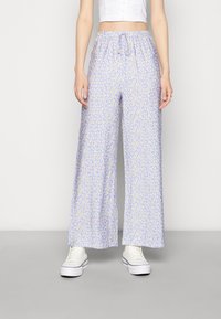 Calças largas azul claro com um padrão floral em branco e amarelo. Apresenta uma cintura elástica com um cordão e são usadas com ténis brancos.
