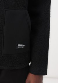 Veste en polaire noire avec une surface texturée et une poche à rabat, ornée d'un label tissé portant l'inscription "REDEFINED REBEL RR-2013."