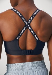 Tamsiai mėlynas sportinis liemenukas su kryžminėmis petnešomis, ant kurių yra "UNDER ARMOUR" prekės ženklas, reguliuojamos pakabos gale ir lygus paviršius. Apačioje matyti šviesūs šortai.