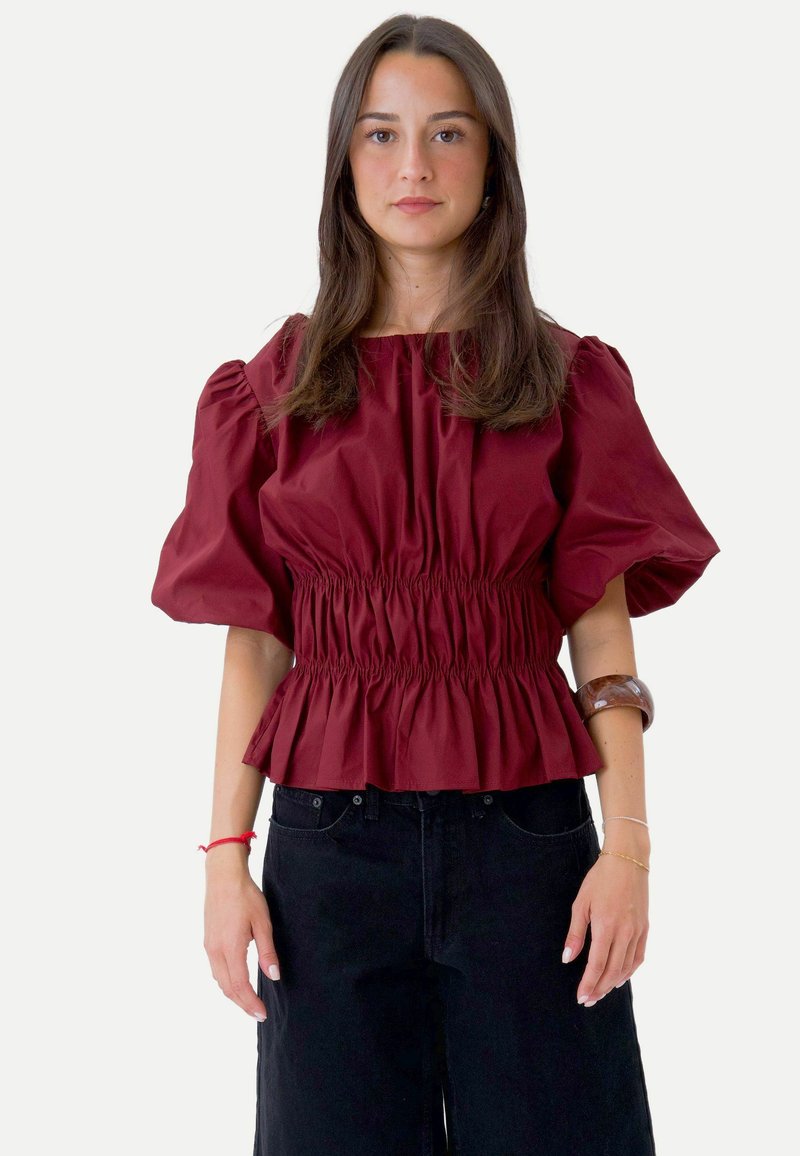 Jeune femme aux longs cheveux bruns portant un chemisier bordeaux à manches bouffantes avec taille froncée et pantalon noir à jambes larges, debout face à l'avant.