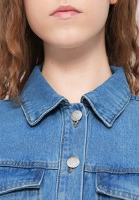 Τζιν μπουφάν σε ανοιχτό μπλε χρώμα, με γιακά που κλείνει με κουμπιά, ασημένια μεταλλικά κουμπιά και δύο τσέπες στο στήθος. Υφή ύφανσης με ορατές ραφές.