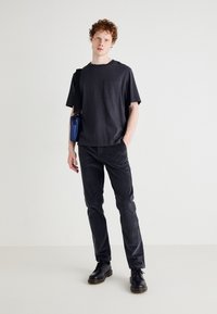 T-shirt noir avec poche, associé à un pantalon gris foncé et des chaussures noires. Un sac bleu est porté en bandoulière. Fond uni.