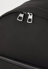 Zaino in tessuto nero con una texture liscia, caratterizzato da doppie zip argentate e un design curvo nella parte superiore. Le cuciture sono ordinate e coerenti.