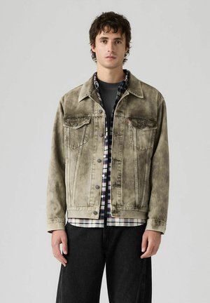 Junger Mann mit kurzen dunklen Haaren, der eine ausgewaschene beigefarbene Jeansjacke über einem karierten Hemd und schwarzen Hosen trägt, stehend vor einfarbigem Hintergrund.