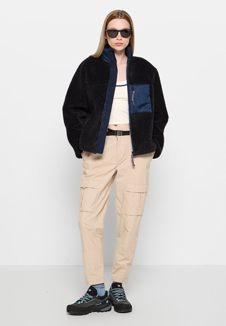 Schwarze Fleecejacke mit einem navyblauen Nylon-Vorderteil, Reißverschluss und Tasche. Beigefarbene Cargohose mit mehreren Taschen. Schwarze Schuhe mit blauen Schnürsenkeln.