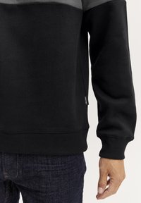 Zwarte sweatshirt met grijze bovenkant, geribbelde mouwen en onderkant. De stof lijkt zacht met een label aan de onderzijde links. G gedragen met een donkere spijkerbroek.