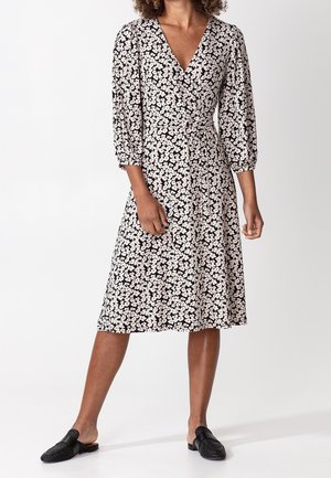 Kwiatowa sukienka kopertowa z głębokim dekoltem w serek, rękawami do połowy przedramienia, czarnym podkładem, różowymi i białymi kwiatami oraz wiązaniem w talii. Dopełniona czarnymi wsuwanymi butami.